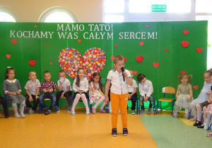 Chłopiec trzyma w ręku mikrofon, recytuje wiersz, w tle siedzą dzieci w kręgu.